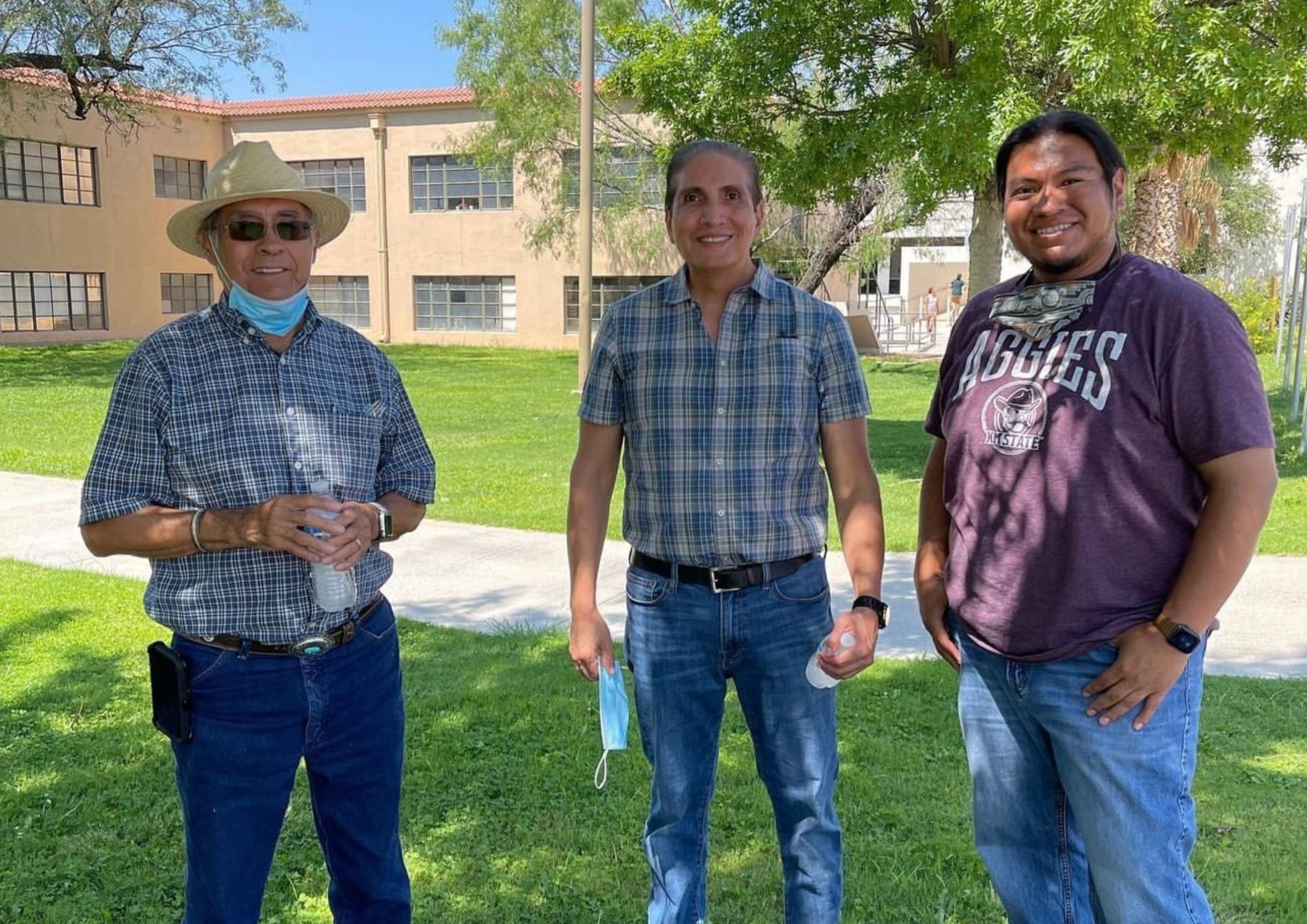 3 Gererations of AIP Directors. The image depicts three men standing outdoors on a green lawn with a large building in the background. They appear to be enjoying a sunny day, as shadows on the grass indicate bright sunlight.  Man on the left: He is wearing a straw hat, sunglasses, a blue plaid shirt, blue jeans, and gray shoes. A light blue face mask hangs below his chin, and he holds a small water bottle and paper cup in his hands. His cell phone is clipped to his belt. Man in the middle: He is wearing a blue and green plaid short-sleeved shirt, blue jeans, and black shoes. He holds a white face mask in his left hand and a water bottle in his right hand. Man on the right: He is wearing a maroon t-shirt with "AGGIES" and a logo printed on it, blue jeans, and black shoes. He has a black watch on his left wrist and a face mask around his neck. In the background, there is a sandy-colored building with many windows. Trees with lush green foliage are also visible, providing shade to some areas of the scene. The sky is clear and blue, indicating pleasant weather.