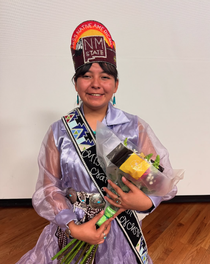 Ms Native American NMSU. Woman wearing traditional regalia, a crown with "MISS NATIVE AMERICAN" and "NM STATE," and holding a bouquet.